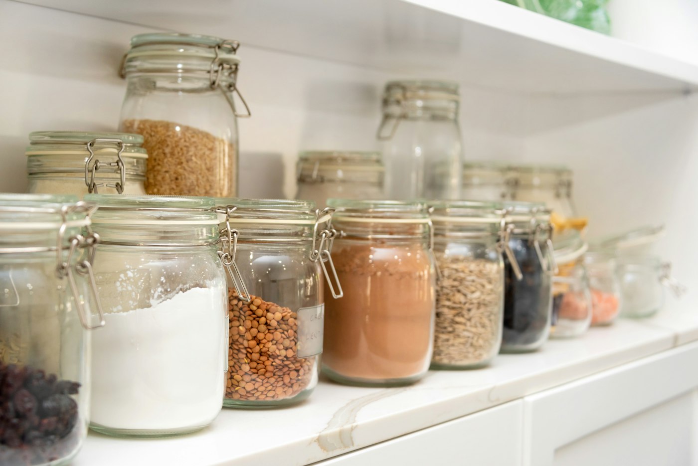 Row of glass jars with clip-top lids holding flour, lentils, cocoa and seeds on a white shelf